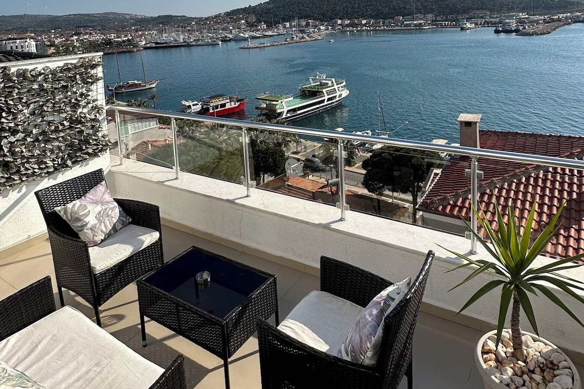 Una terraza con mobiliario de mimbre, una planta en maceta y vistas a un puerto deportivo con barcos y una colina distante.