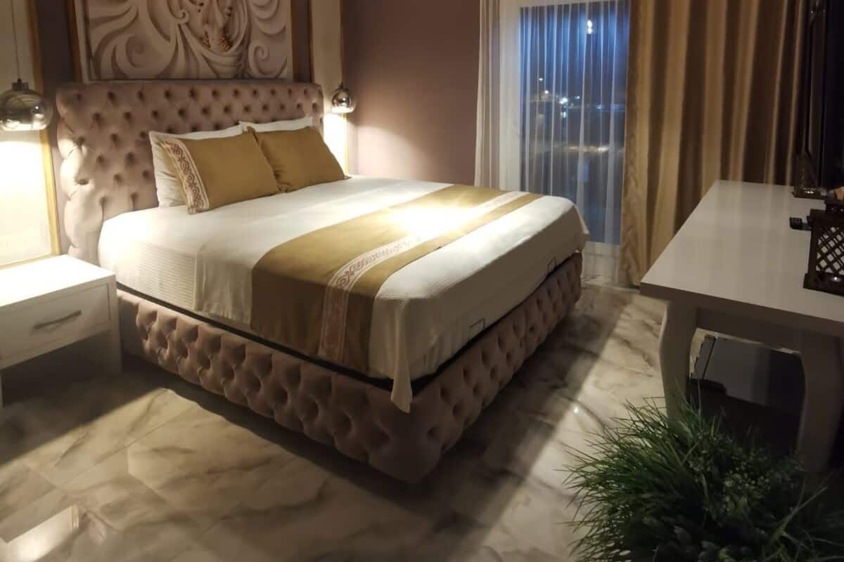 Un dormitorio con cabecera capitonada, una cama grande con colcha beige y blanca, una mesita de noche, una lámpara colgante y vistas de la ciudad a través de cortinas.