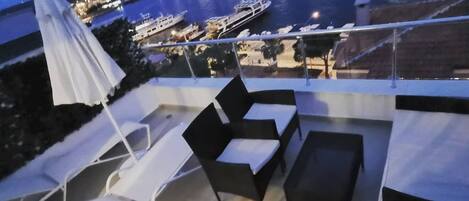 Un balcón con mobiliario exterior, una sombrilla blanca y vista a un puerto deportivo al anochecer.