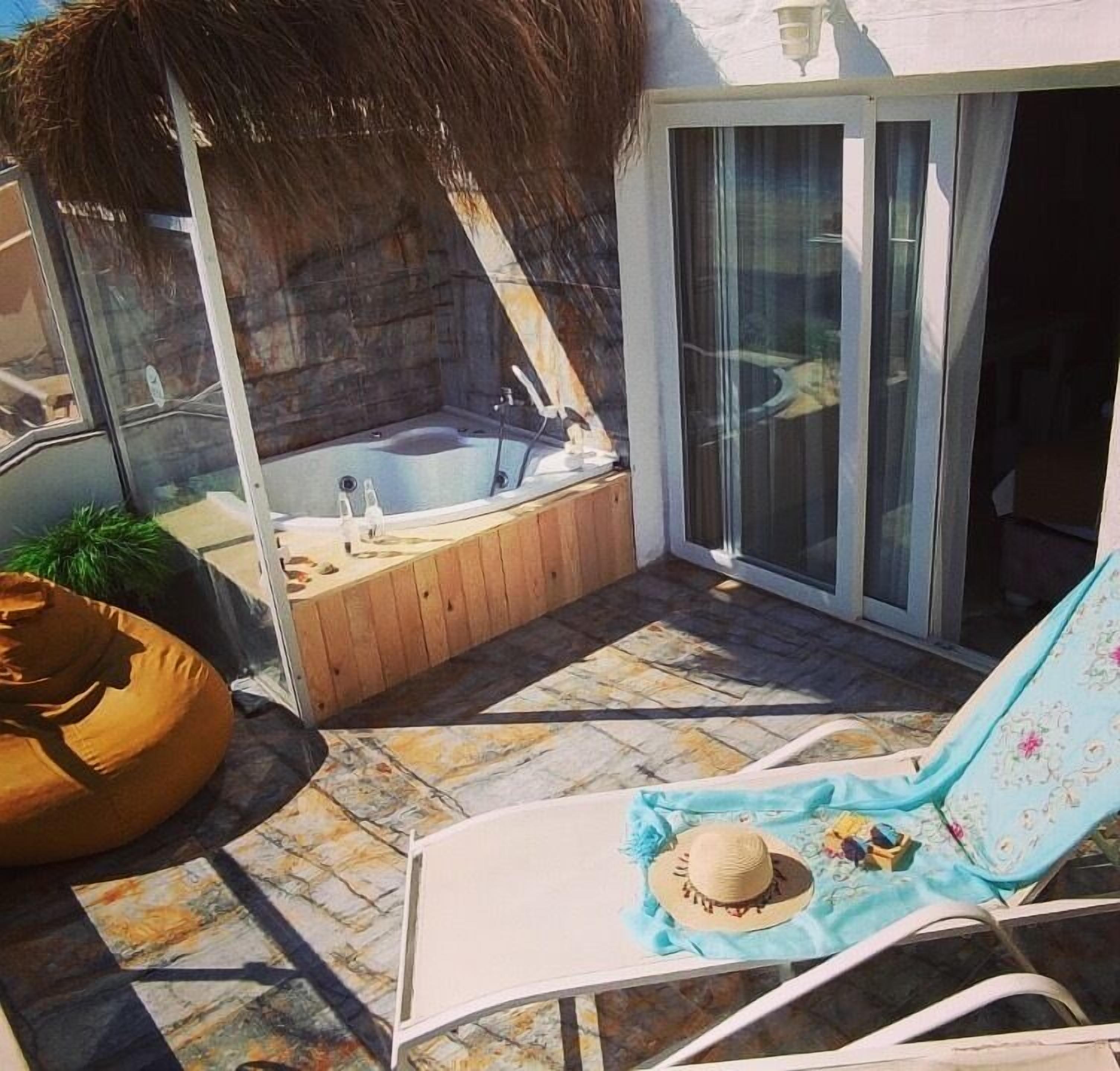 A sunlit patio with a hot tub, a lounge chair, and a thatched roof structure.