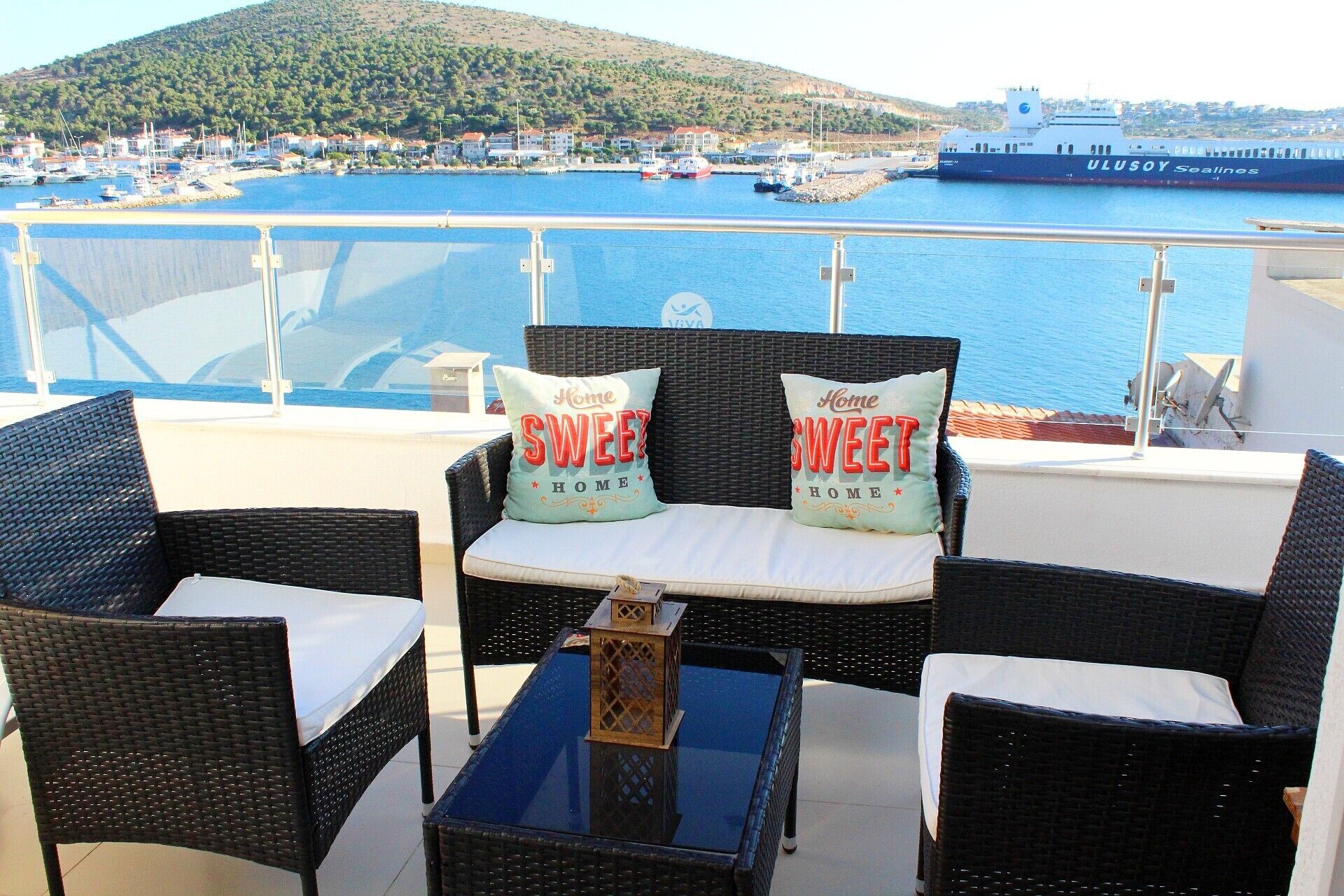 A balcony with wicker furniture, a glass table, and a view of a harbor with boats and a ship.