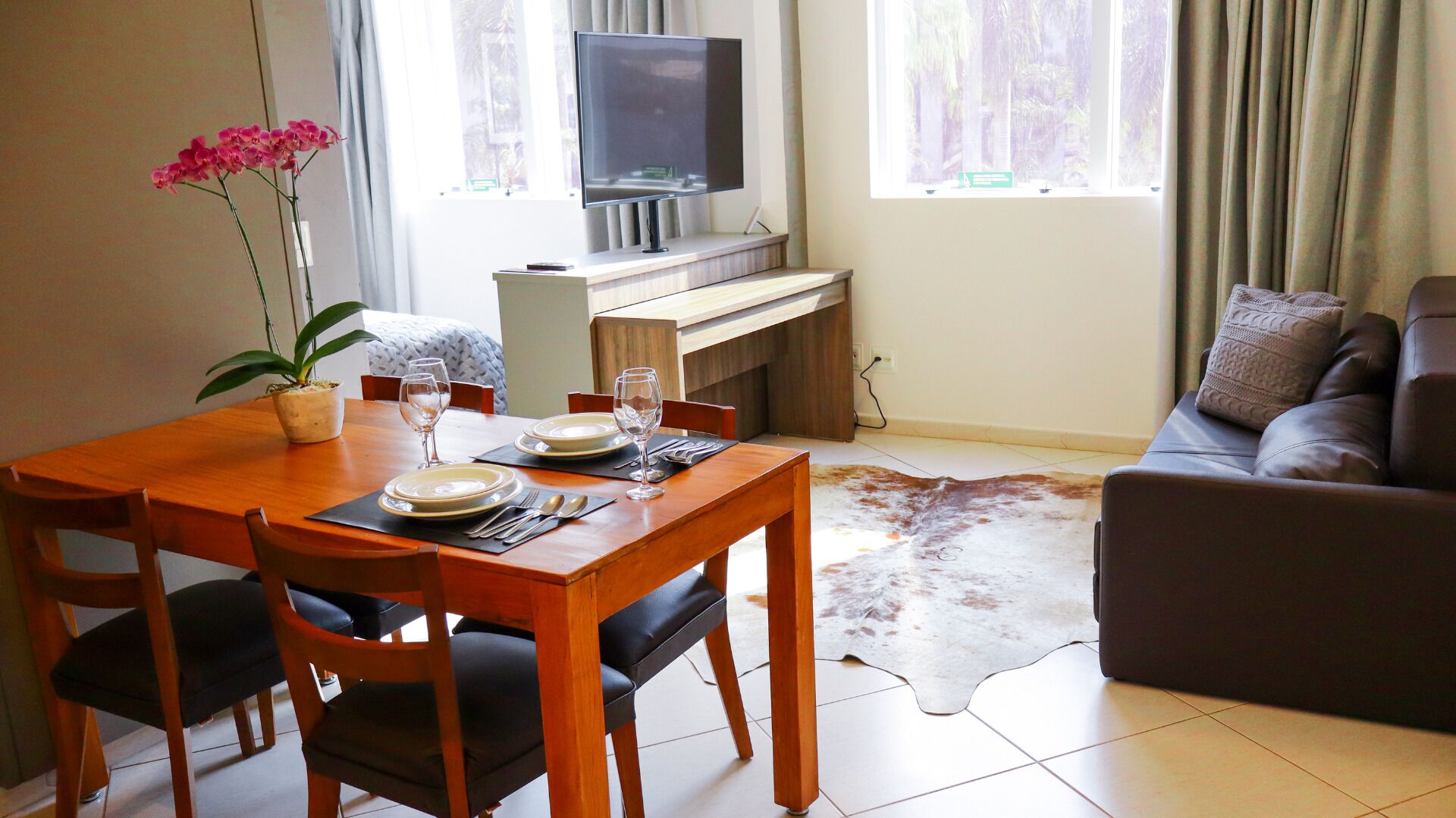 A dining area with a wooden table set for two, a television on a stand, and a leather sofa in the living area.