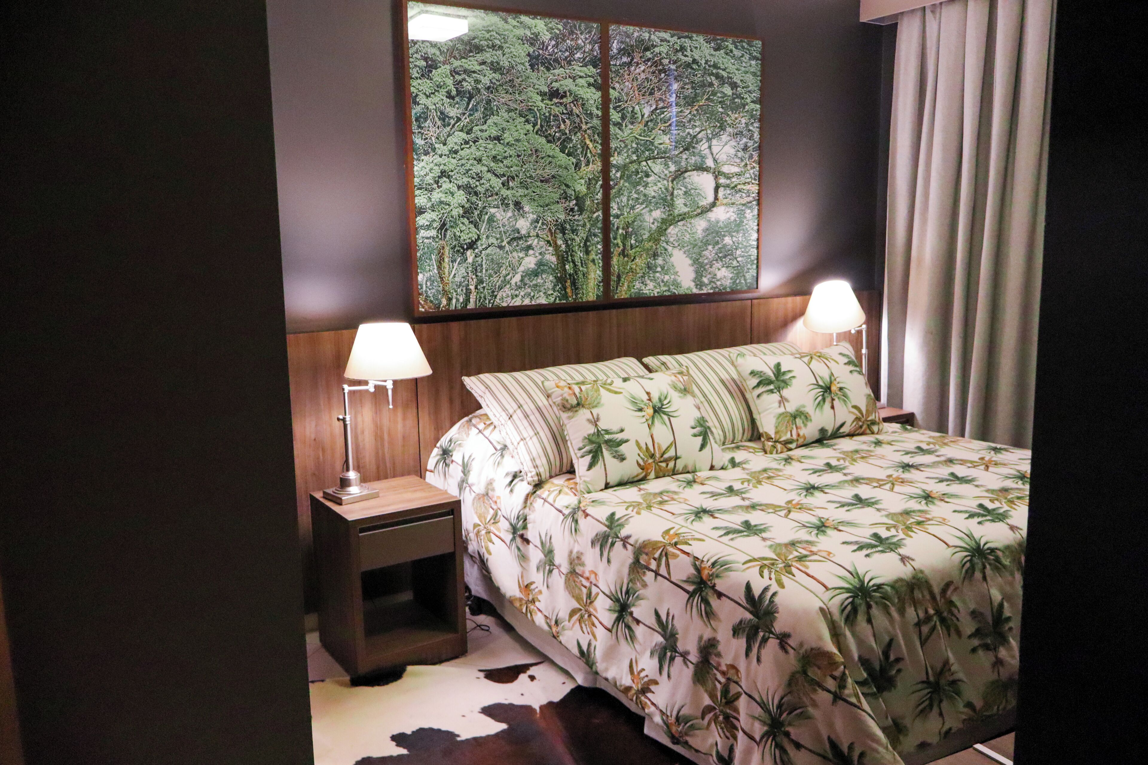 Un dormitorio con cama, dos lámparas de noche, una mesita de noche de madera, un cuadro grande con un paisaje de árboles y una ventana con cortinas.