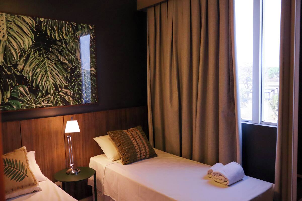 A hotel room with a bed, a nightstand, a lamp, a window with curtains, and a wall with a tropical leaf pattern.