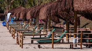 A beach with rows of wooden lounge chairs under thatched umbrellas, a sandy beach, and palm trees in the background. - Barretos Park Hotel (Barretos)