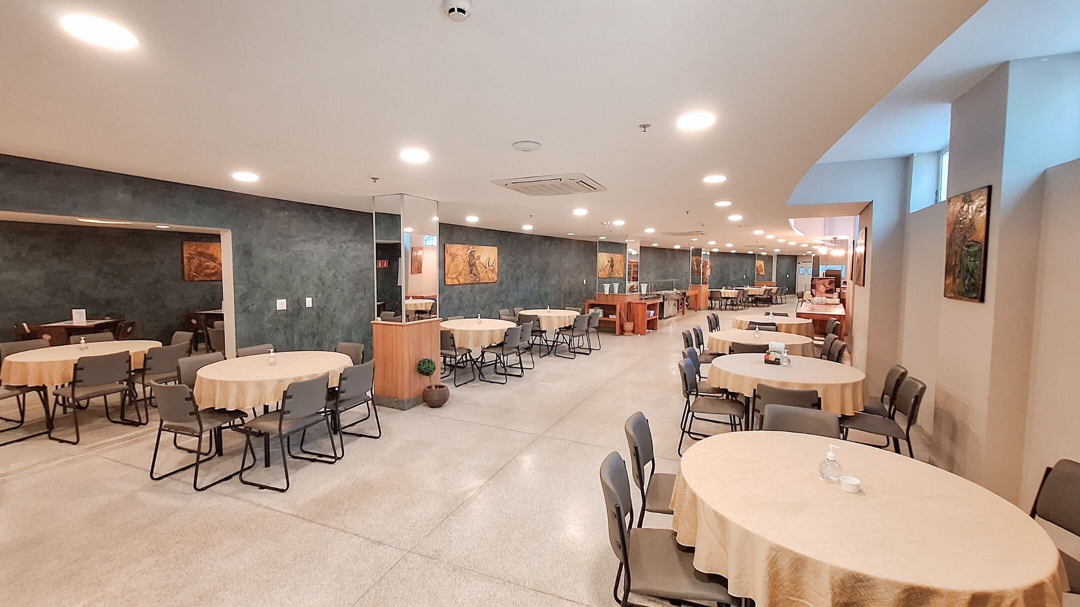 A spacious dining area with multiple round tables and chairs, a counter with a cash register, and artwork on the walls.