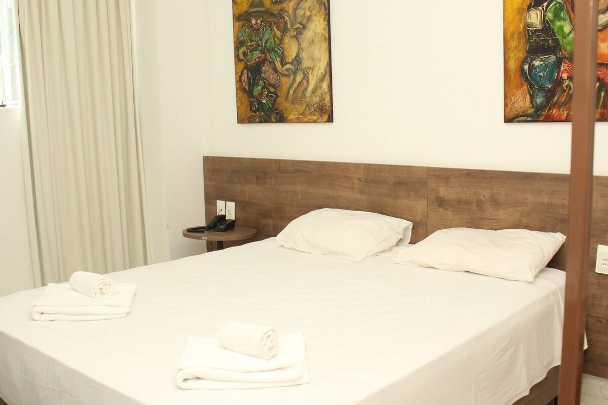 A hotel room with a wooden headboard, a bed with white linens, two rolled towels, a bedside table with a phone, and two paintings on the wall.