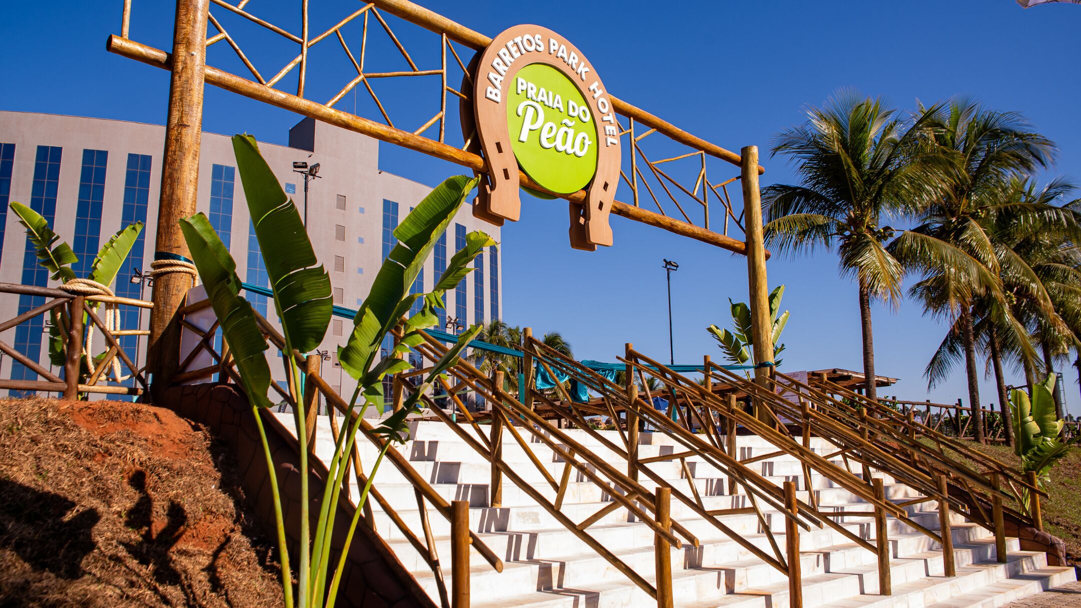 Entrada de un hotel con un letrero que dice 'Barreitos Park Hotel Praia do Peão', rodeada de palmeras y una escalera.