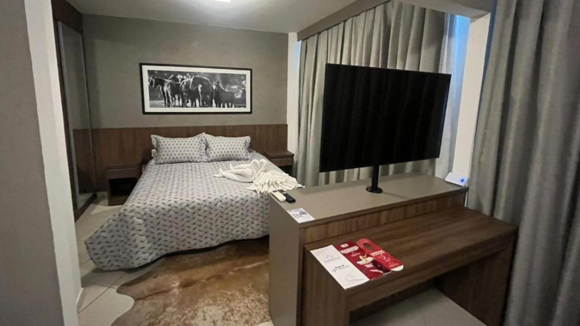A hotel room with a bed, a desk with a TV, a framed picture on the wall, and a window with curtains.