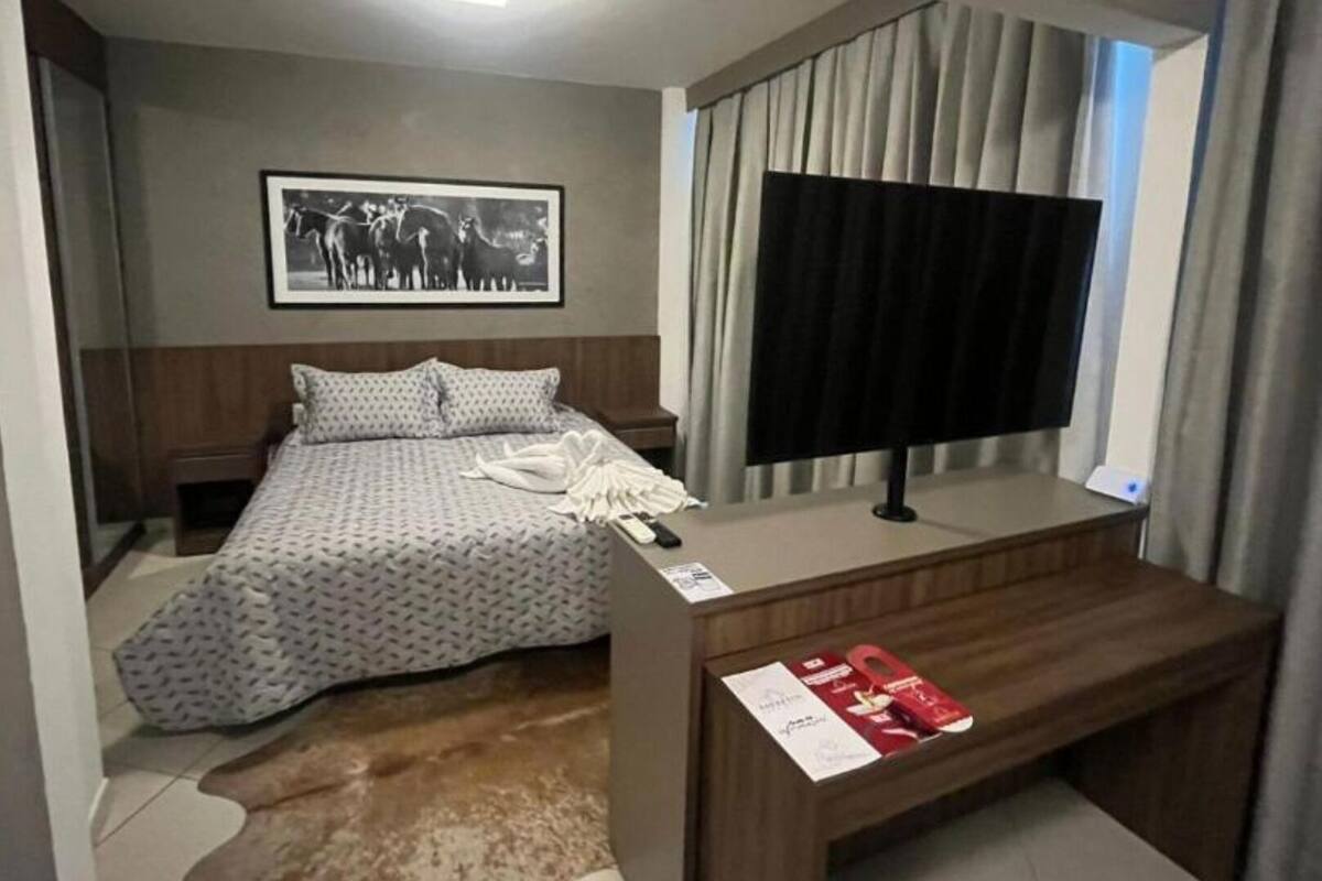 A hotel room with a bed, a desk with a TV, a framed picture on the wall, and a window with curtains.