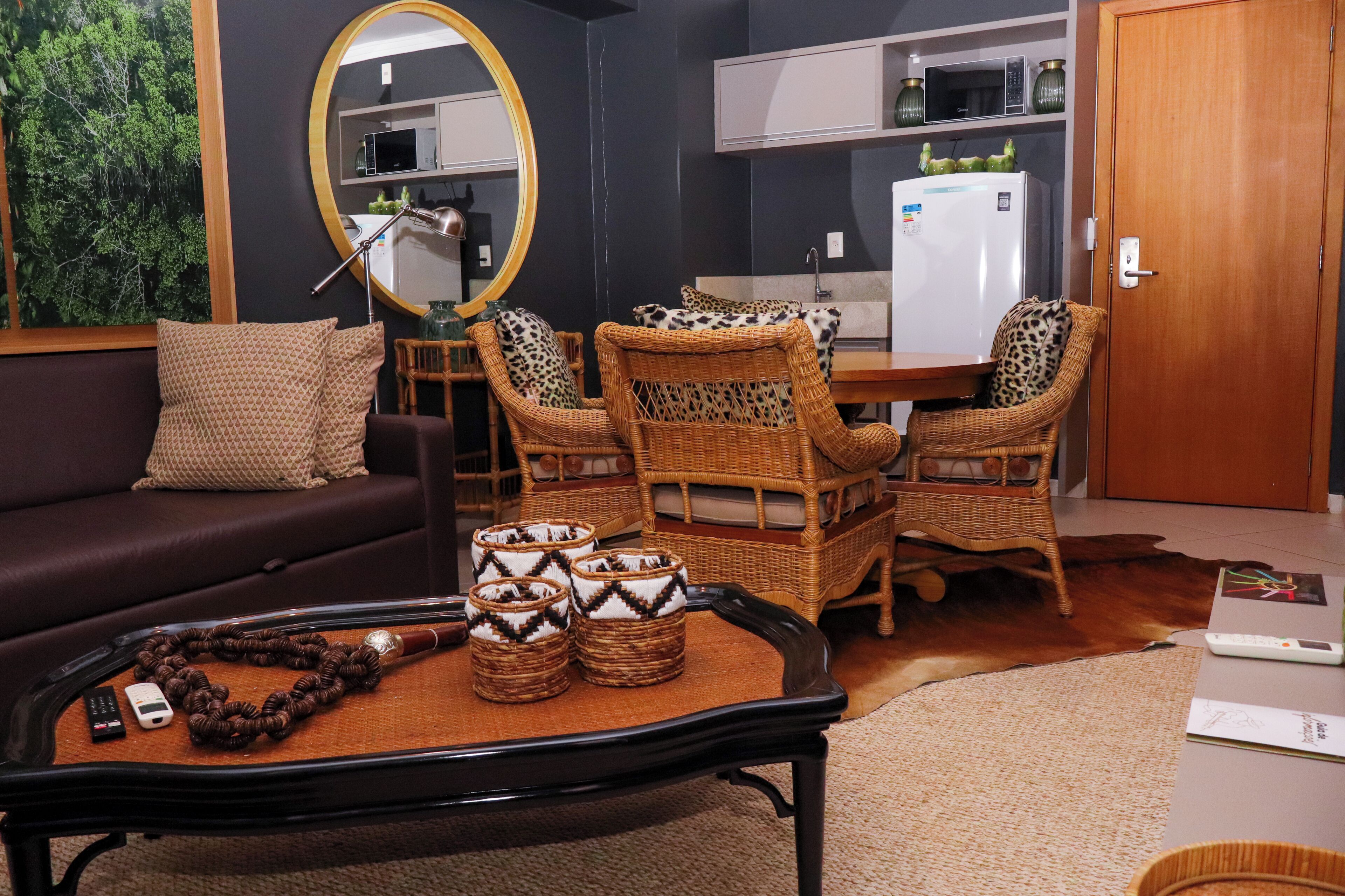 A living room with a sofa, two wicker chairs, a coffee table with decorative items, a mirror, and a kitchen in the background.