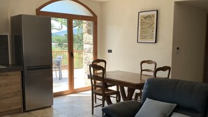 Een woonkamer met een bank, een eettafel met stoelen, een keuken met een koelkast, en een grote schuifpui die uitkomt op een terras.