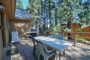 A wooden deck with a table and chairs, a grill, and a view of tall trees. - Rim Drive Lookout (Tahoe Vista)