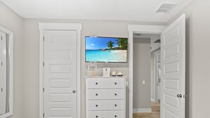 Un couloir avec une commode blanche, un téléviseur diffusant une scène de plage et des portes blanches.