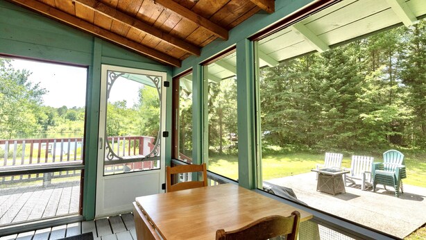 Una terraza de madera con una mesa y sillas, ventanas amplias y vistas a un bosque.
