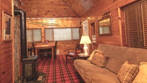 Un acogedor interior de cabaña con alfombra escocesa, paredes de madera, un sofá, una mesa con sillas y una lámpara.
