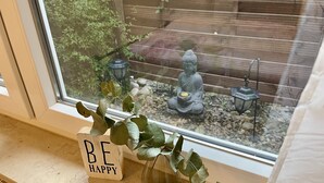 Une fenêtre donnant sur un patio, avec une plante en pot et une pancarte portant l’inscription « SOYEZ HEUREUX » posée sur le rebord de la fenêtre.
