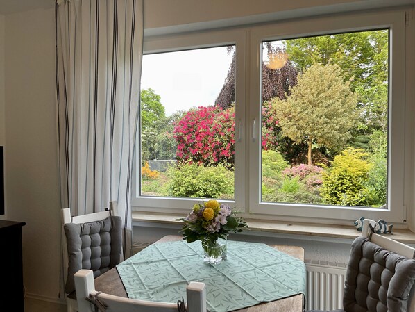 Un comedor con una mesa dispuesta para dos, una ventana con vista a un jardín y un florero con flores sobre la mesa.
