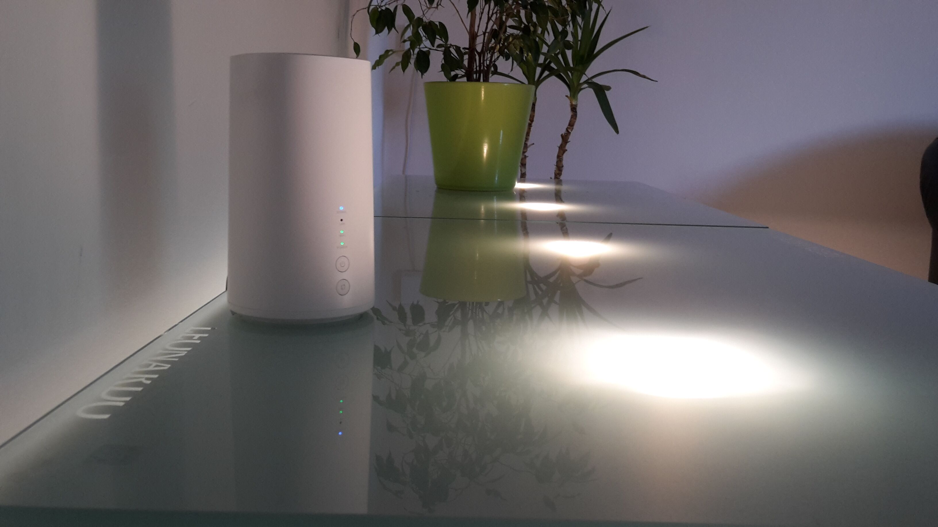 Un dispositivo blanco sobre una mesa de vidrio, con una planta verde en maceta a su lado.