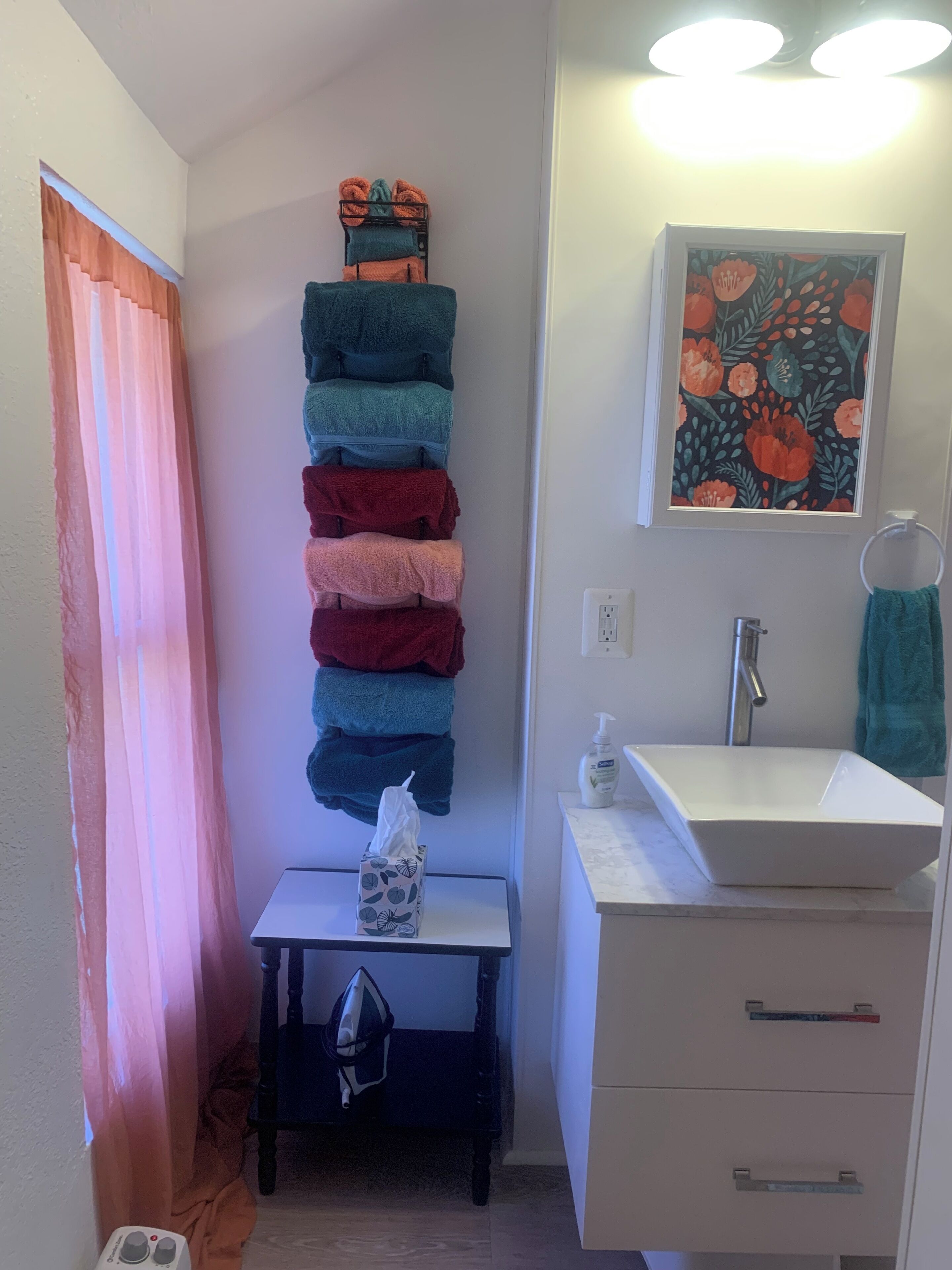 A bathroom with a towel rack holding multiple towels, a sink with a mirror, a framed floral artwork, and a small table with a tissue box and a bag.