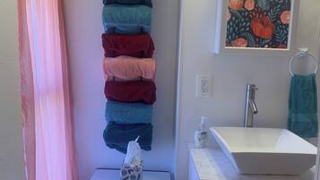 A bathroom with a towel rack holding multiple towels, a sink with a mirror, a framed floral artwork, and a small table with a tissue box and a bag.
