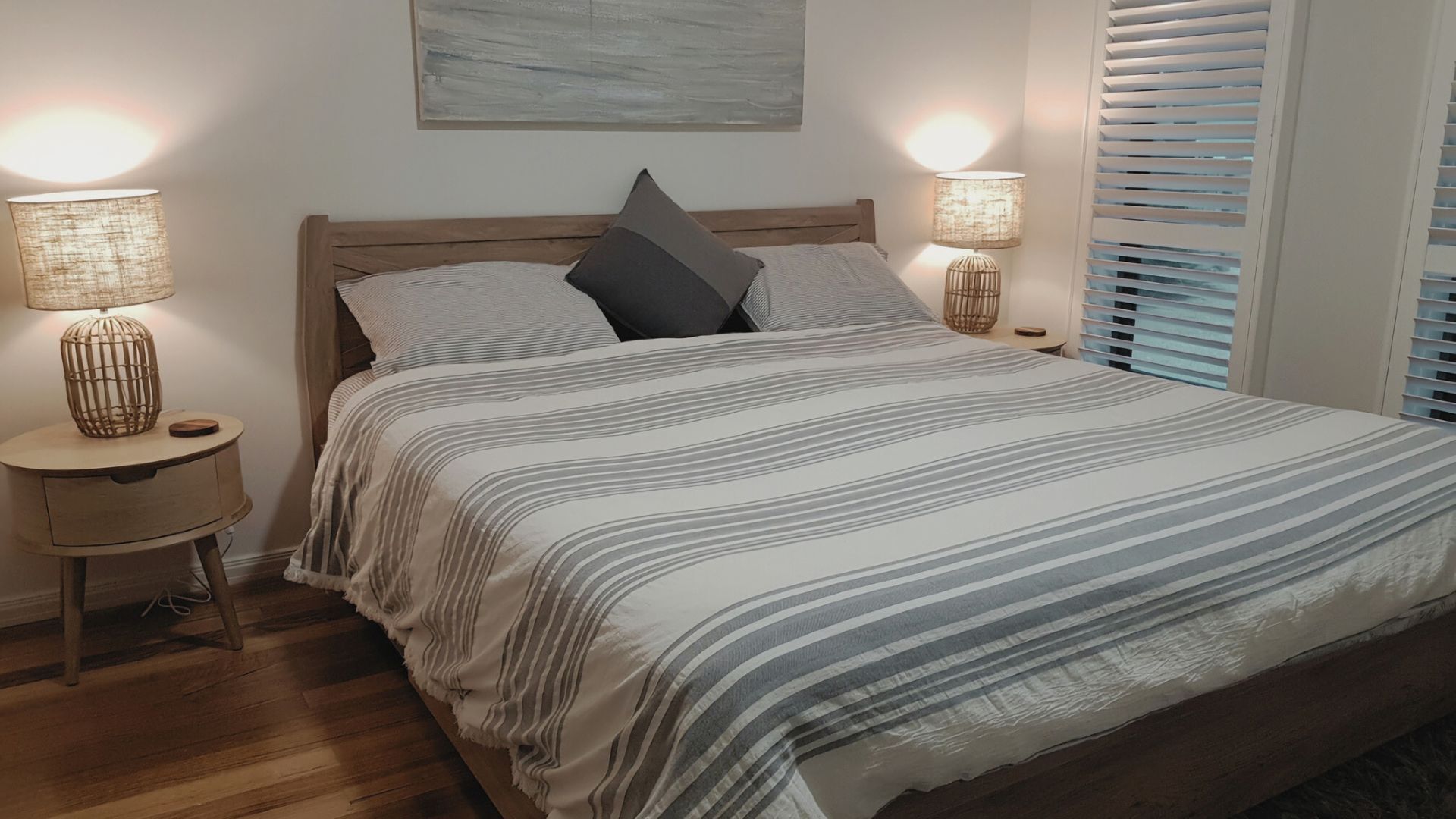 A neatly made bed with striped bedding, two bedside tables with lamps, and a window with blinds.