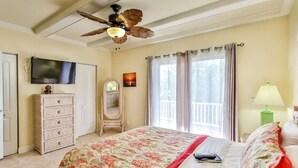 Ein Schlafzimmer mit einem Bett, einem Deckenventilator, einem Fernseher, einer Kommode, einem Spiegel und einem Fenster mit Vorhängen.