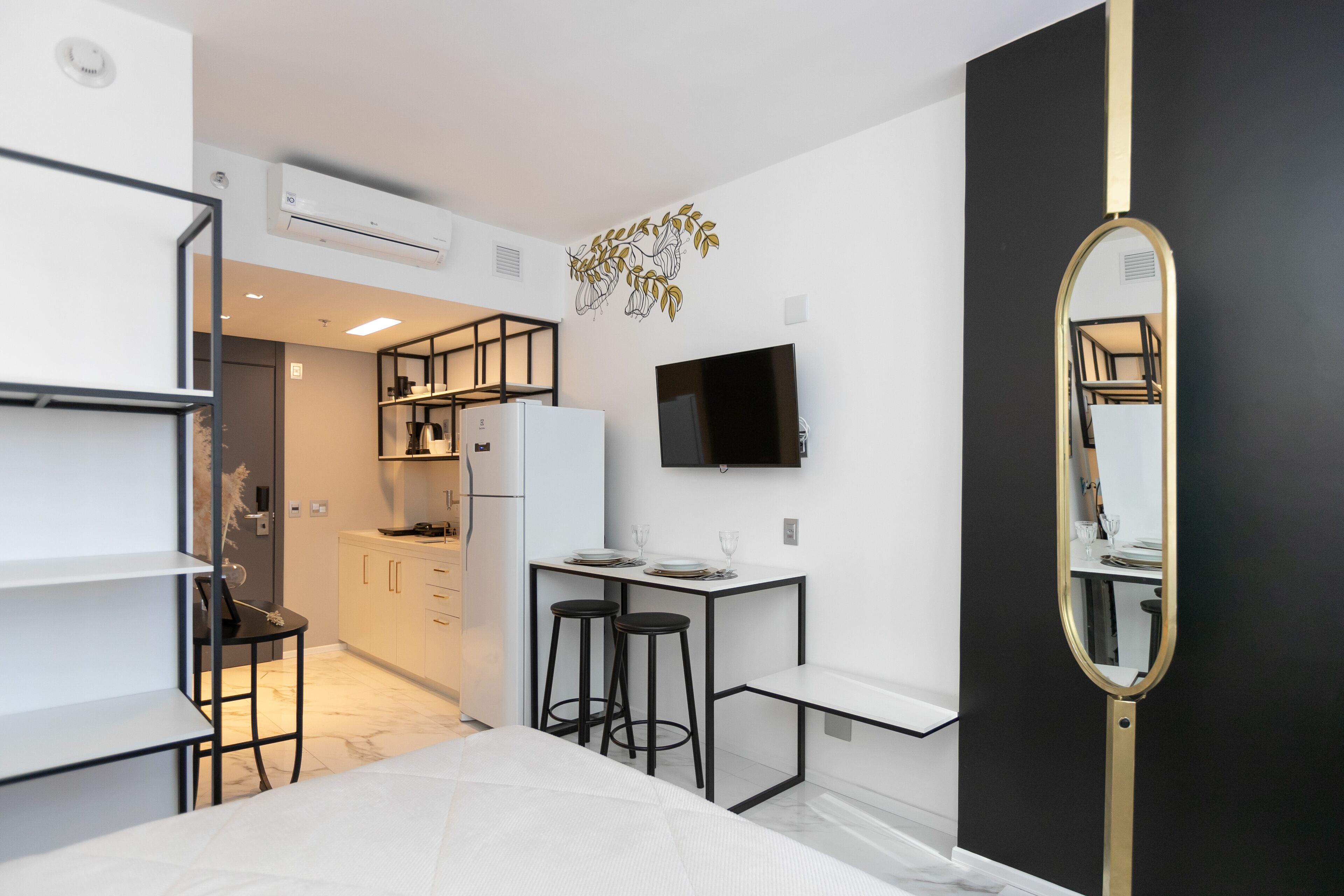 A modern hotel room with a bed, a desk, two bar stools, a flat-screen TV, and a large mirror.