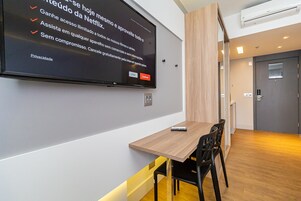 Una habitación de hotel moderna con un televisor de pantalla plana grande montado en la pared mostrando un anuncio de Netflix. Hay una mesa de madera con dos sillas, un ventanal pequeño y una puerta al fondo.