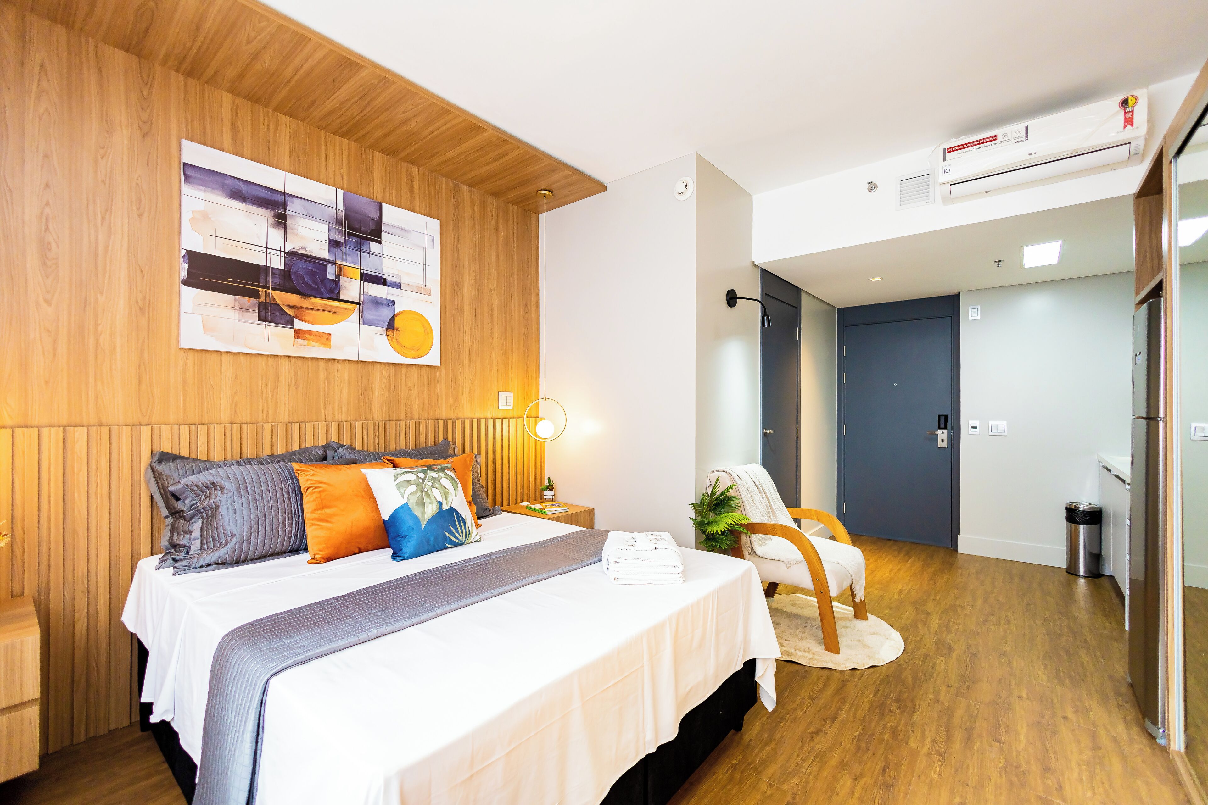 A modern hotel room with a wooden headboard, a large abstract painting, a neatly made bed with multiple pillows, a small bedside table with a lamp, and a view into the adjacent bathroom.