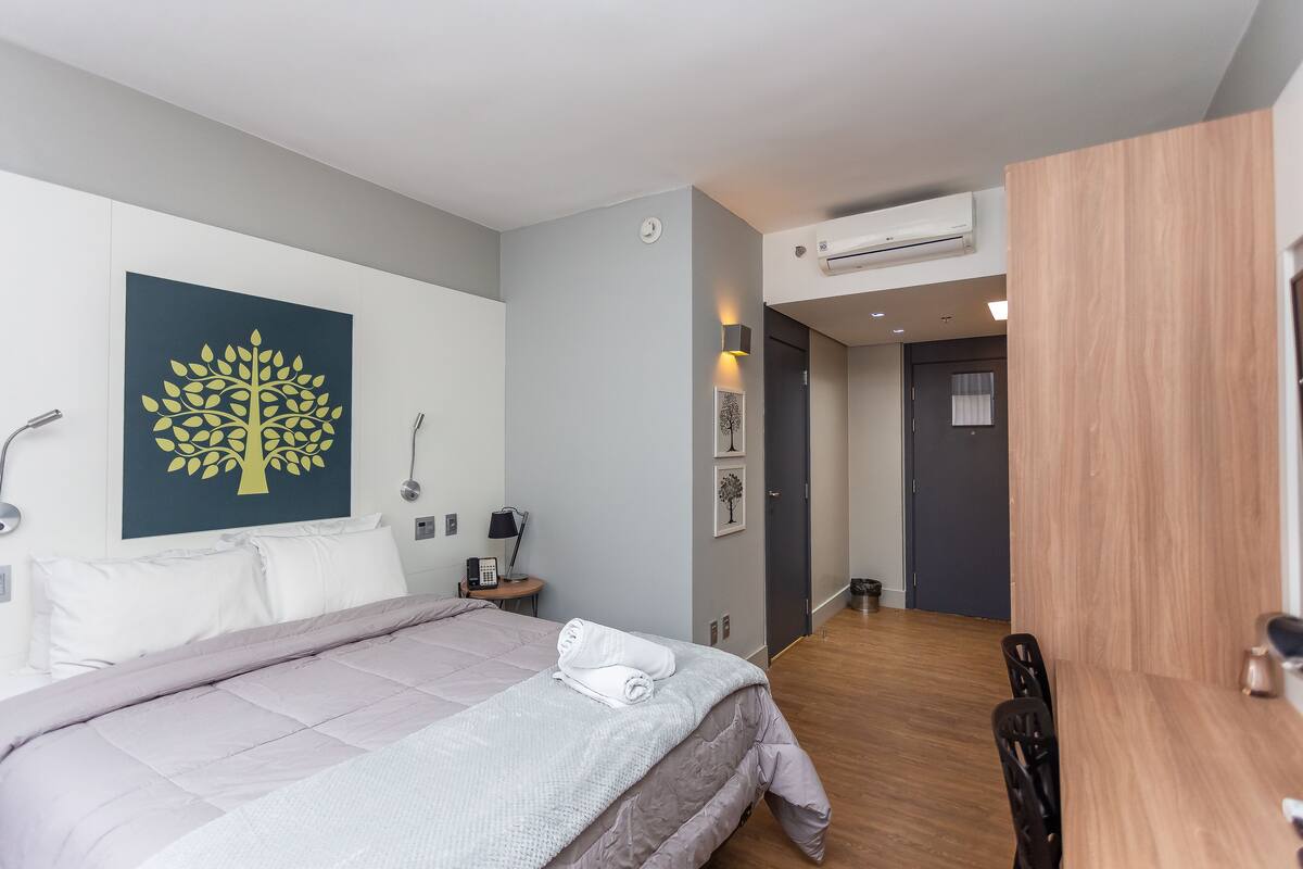 Une chambre d’hôtel moderne avec un grand lit, une œuvre d’art sur le thème des arbres accrochée au mur, une petite table de chevet avec une lampe et une table à manger en bois.
