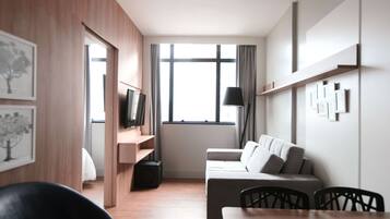 A modern hotel room with a wooden floor, a sofa, a dining table, and a TV.