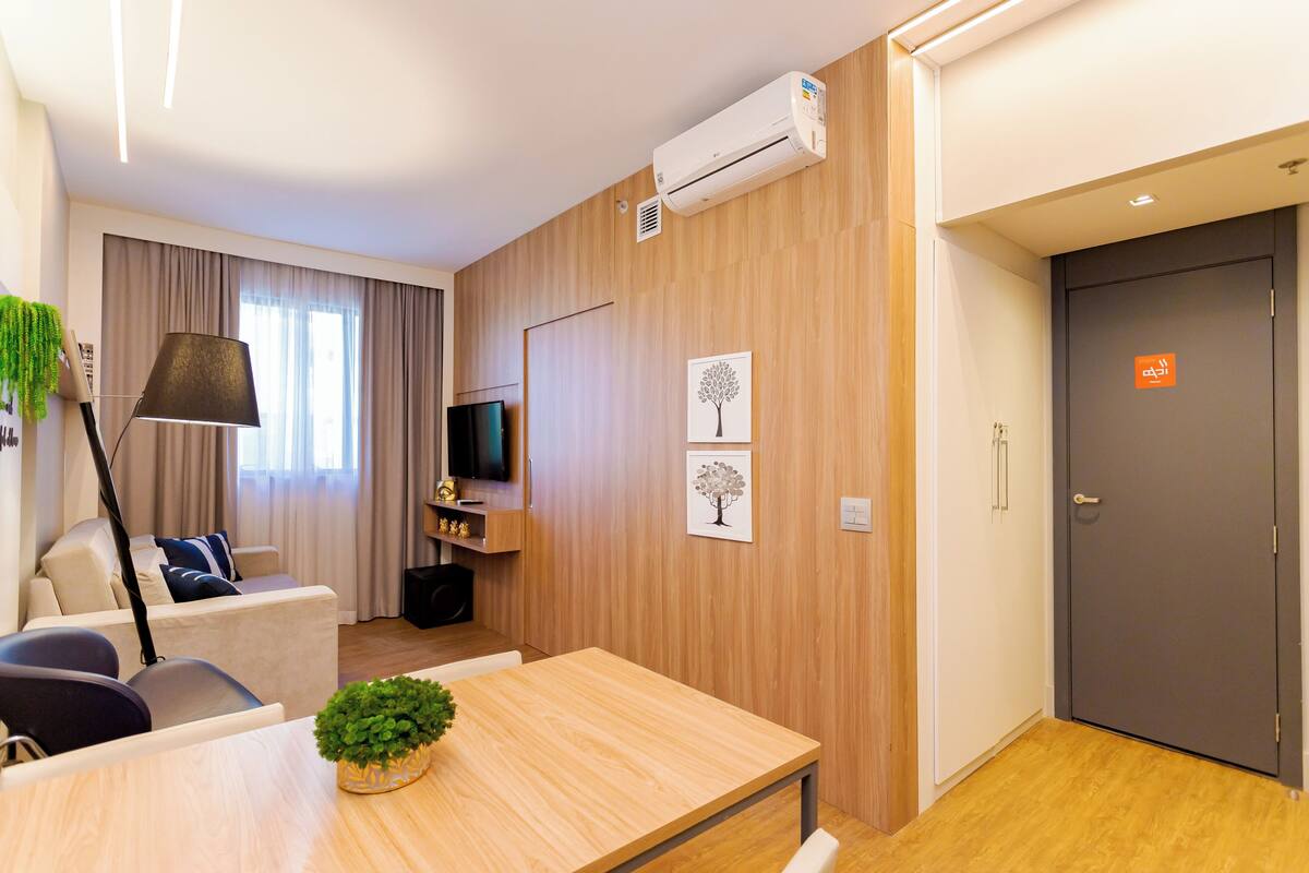 Une chambre d’hôtel moderne avec un plancher en bois, une table à manger agrémentée d’une plante, un canapé avec un téléviseur et une porte sur laquelle est apposé un panneau indiquant la sortie.