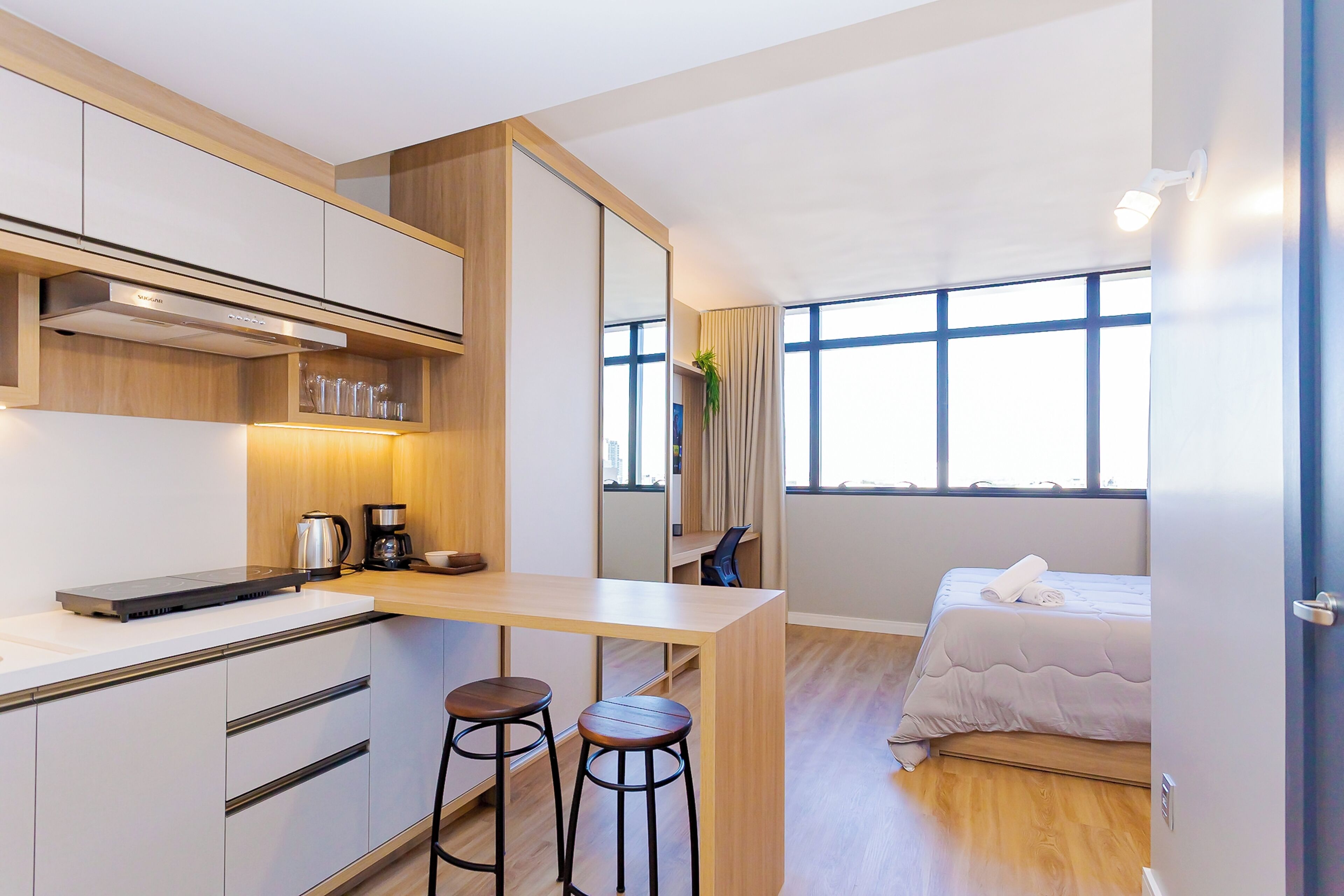 A compact hotel room with a kitchenette, a bed with a white duvet, and a large window providing natural light.