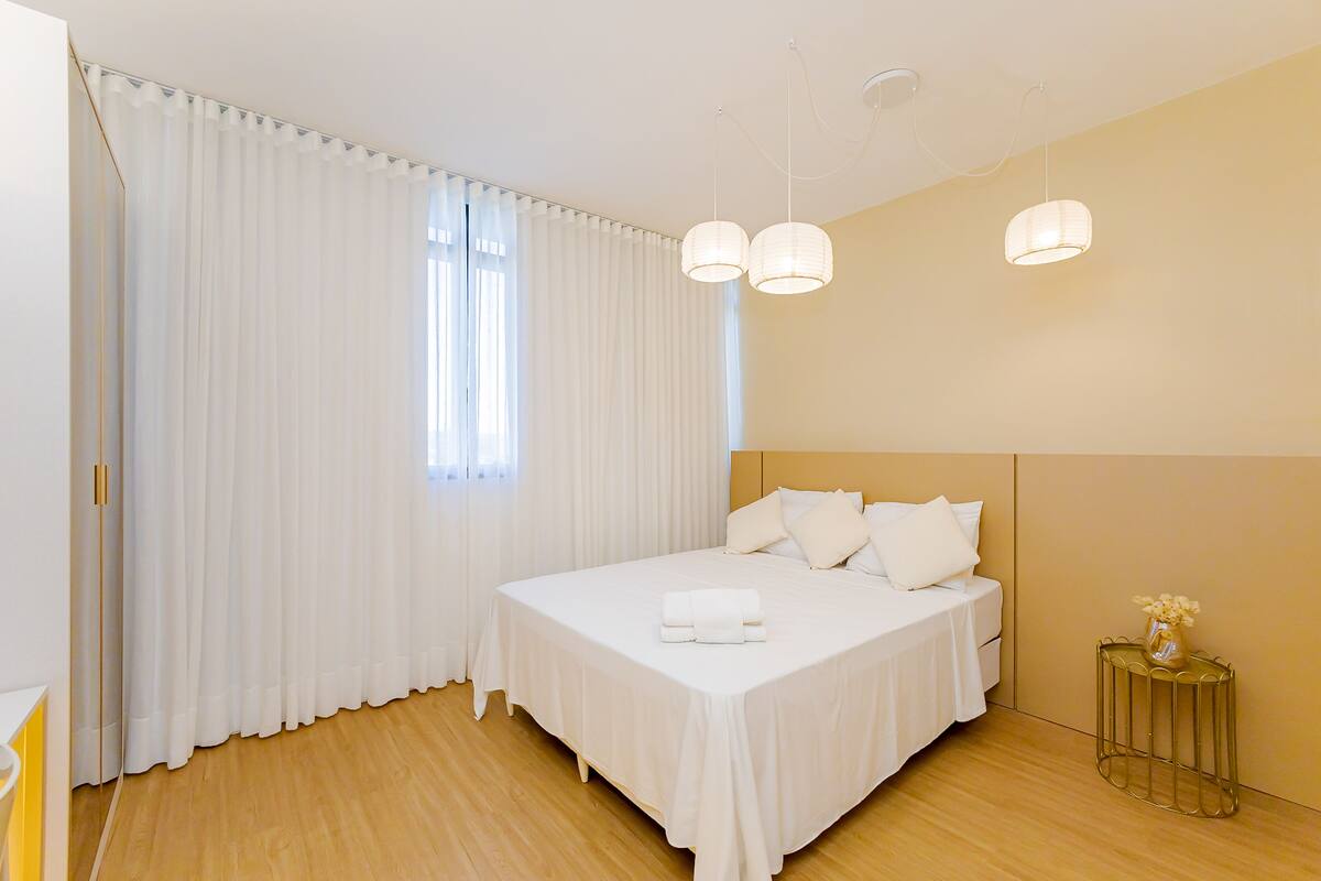 Une chambre d’hôtel moderne avec un grand lit, des rideaux blancs et une petite table d’appoint sur laquelle est posé un vase.
