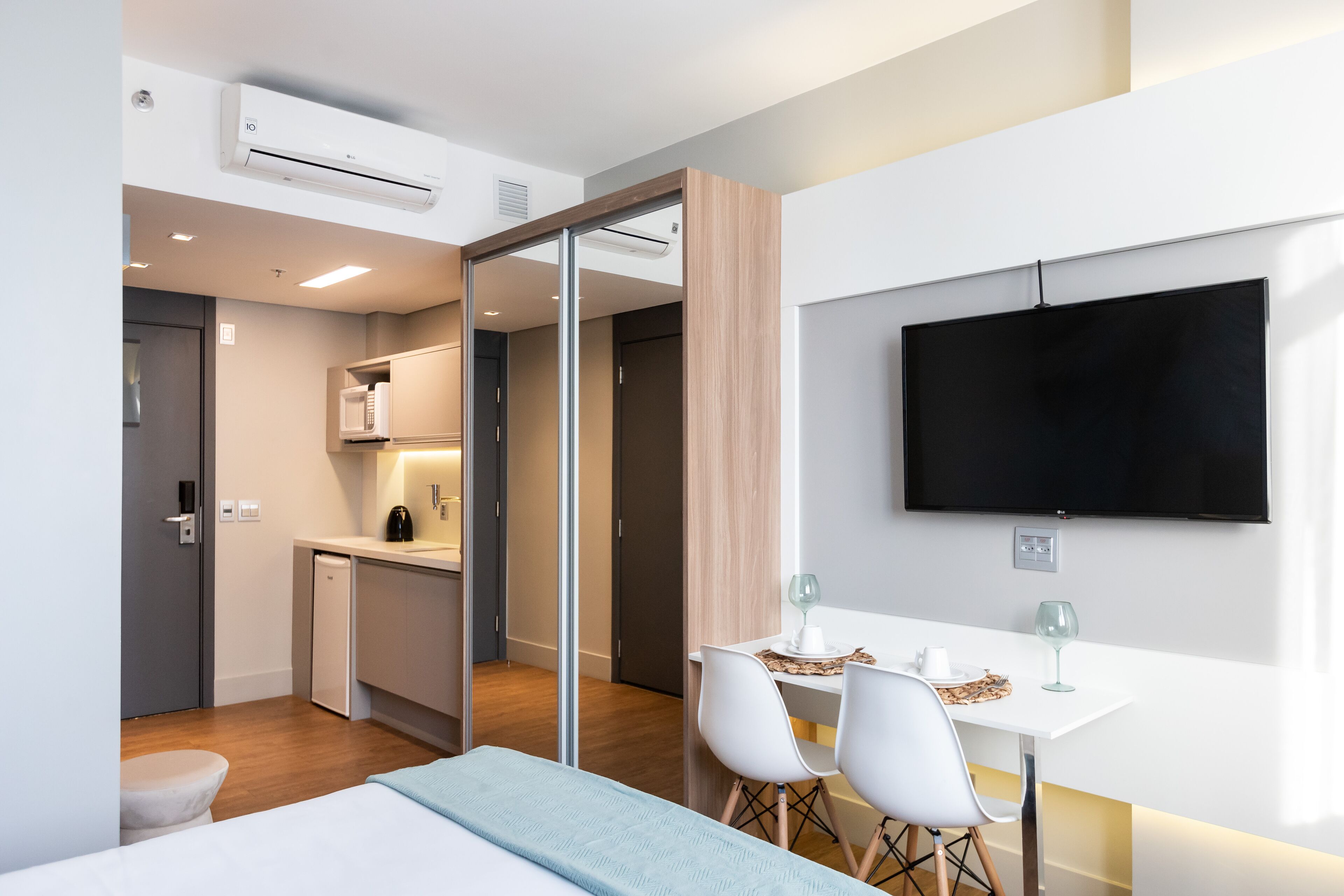 A modern hotel room with a bed, a desk with two chairs, a TV, a kitchen area, and a sliding door.