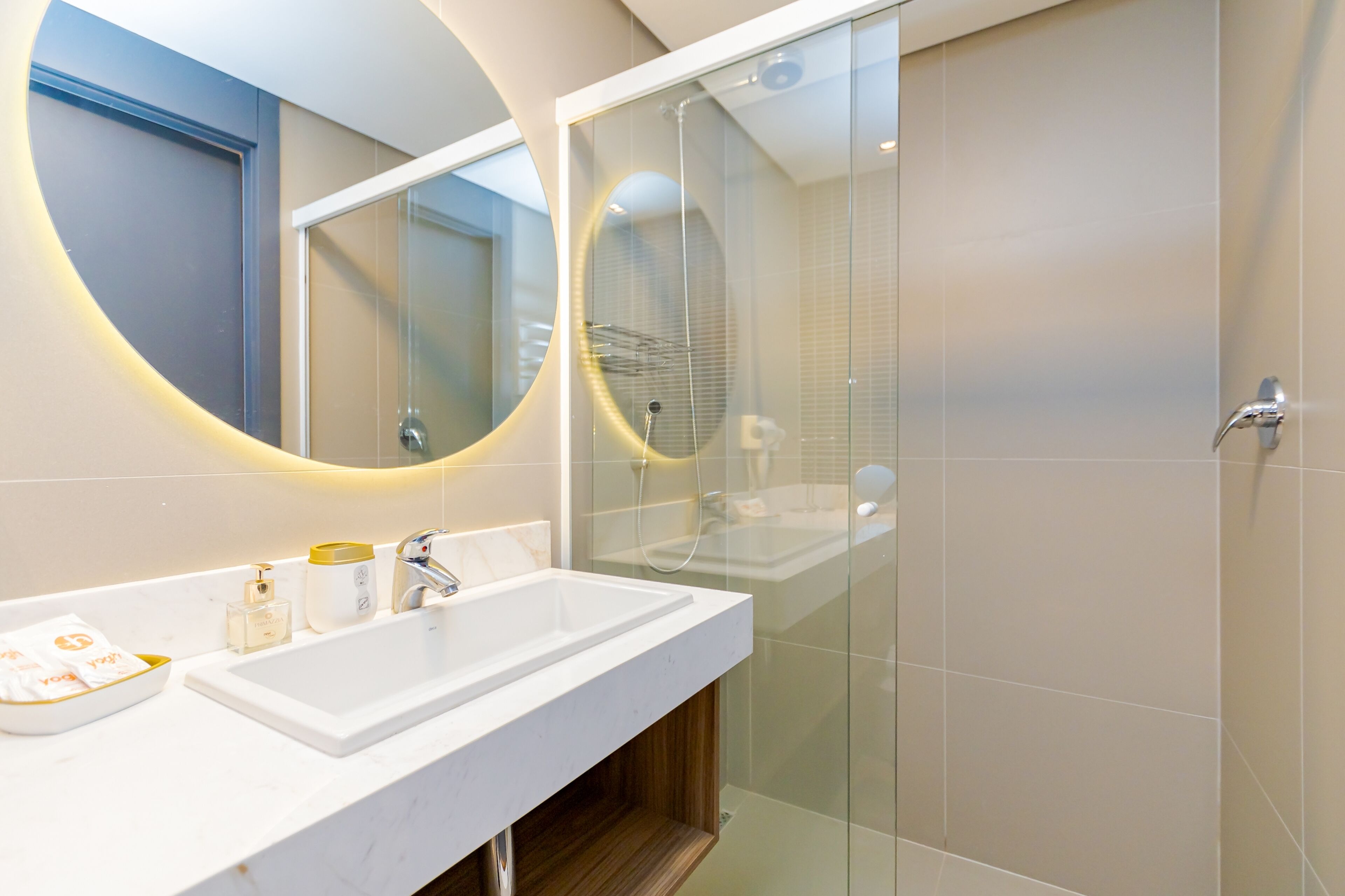 A modern bathroom with a white sink, a glass shower enclosure, and a large mirror with a circular light.