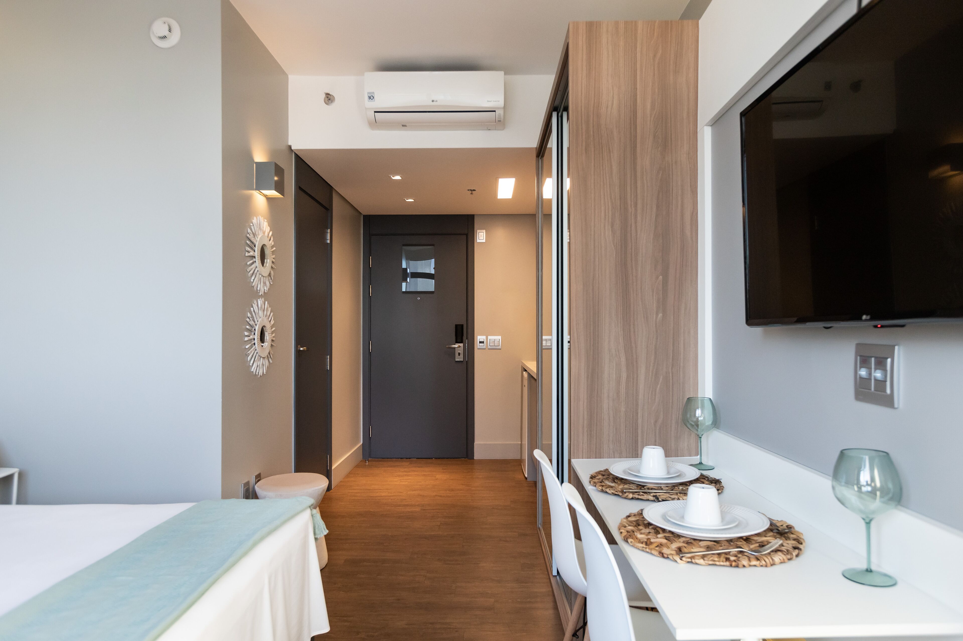 Quarto de hotel moderno com área de refeições, TV fixada na parede e porta para o corredor.