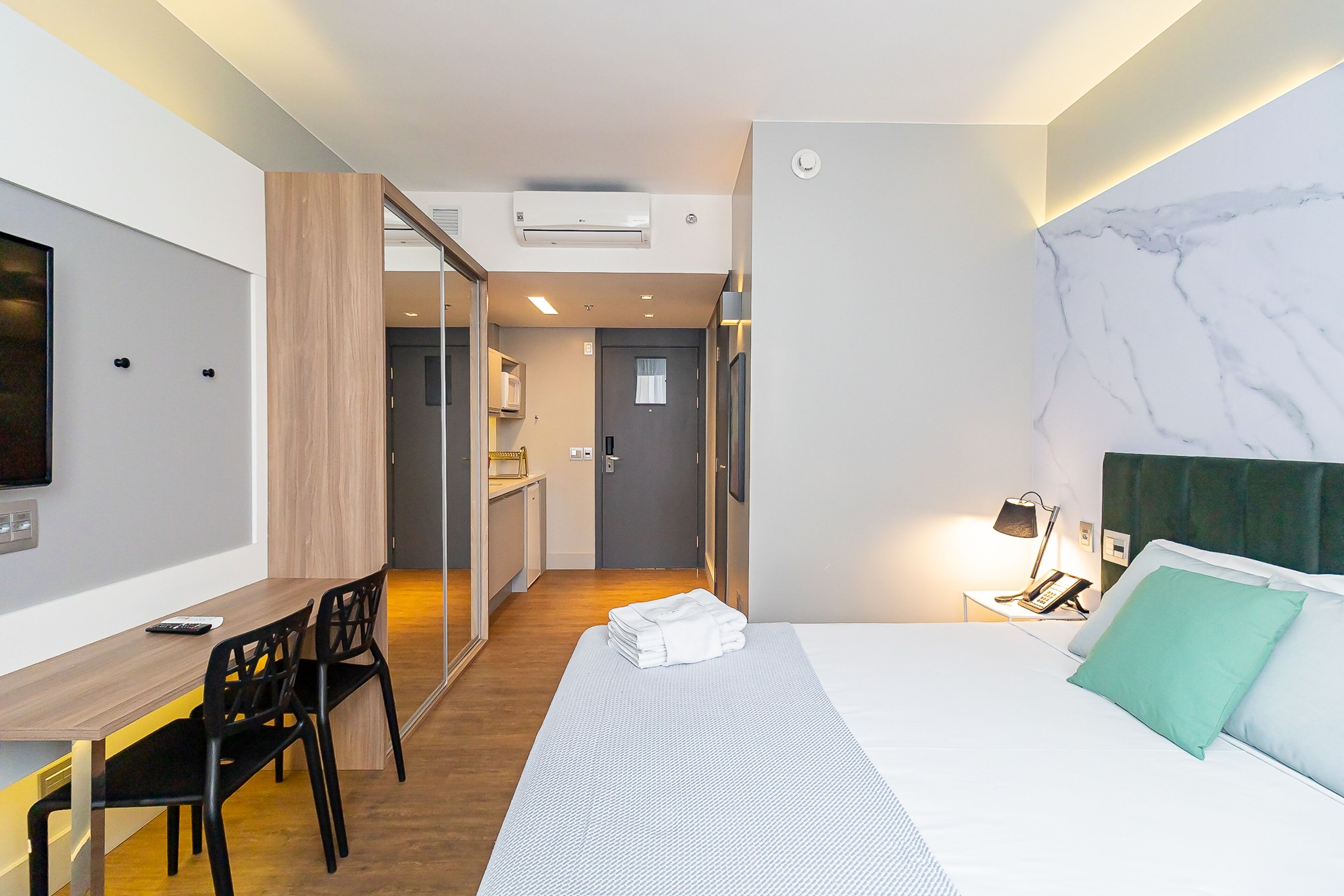 Une chambre d’hôtel comprenant un lit, un bureau, une chaise, une télévision, une table de chevet avec une lampe, et une salle de bain visible à travers une porte en verre.