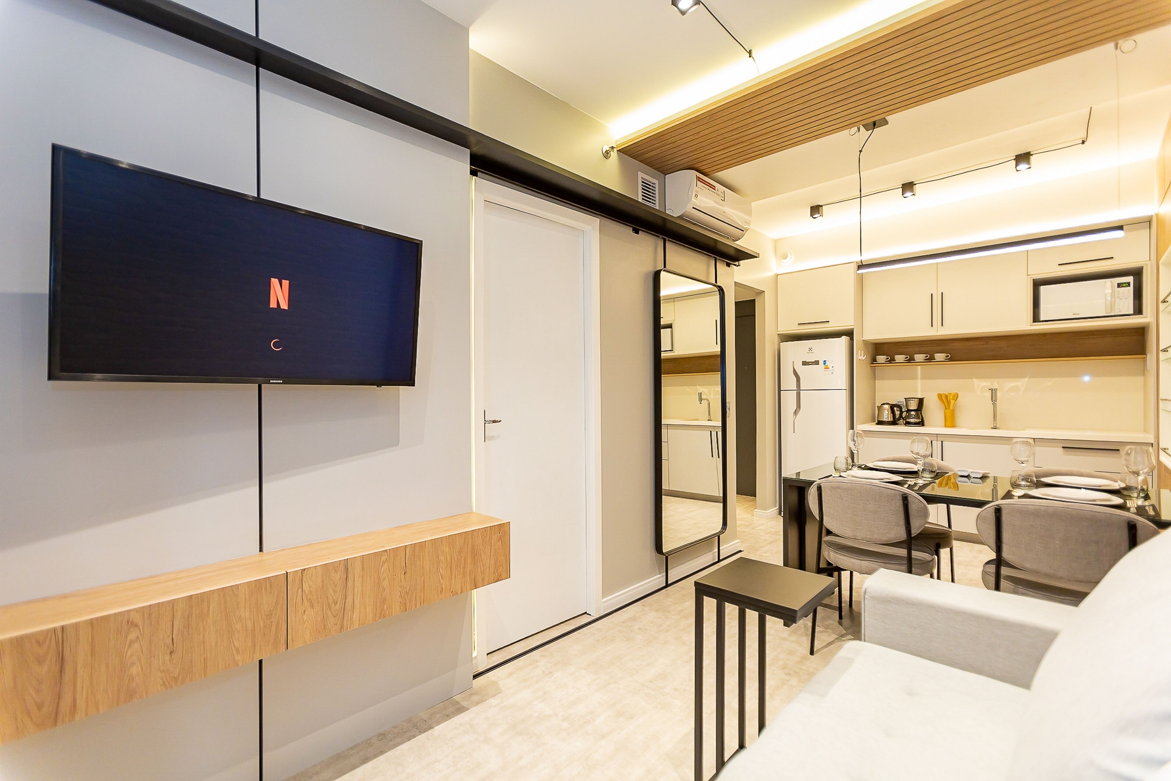 A modern hotel room with a flat-screen TV mounted on the wall, a wooden bench, a dining area with a table and chairs, and a kitchenette with a microwave and coffee maker.
