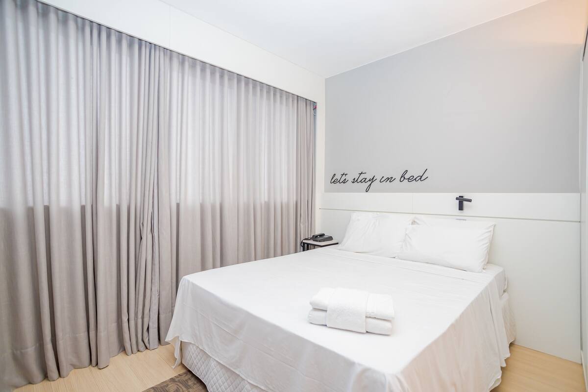 Un lit bien fait, avec du linge de lit blanc et une serviette pliée, un téléphone sur la table de chevet et un mur orné d’une croix et de l’inscription : « restons au lit ».