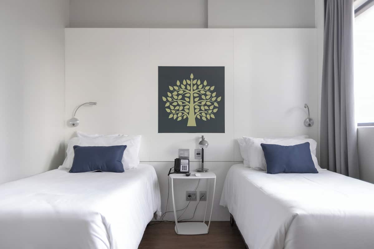 Une chambre d’hôtel avec deux lits, une table de chevet avec un téléphone et une lampe, et une œuvre d’art représentant un arbre, accrochée au mur.