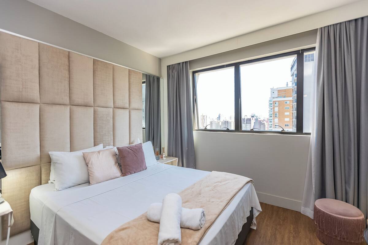 Une chambre d’hôtel avec un grand lit, une vue sur la ville par la fenêtre, une table de chevet et un petit tabouret.