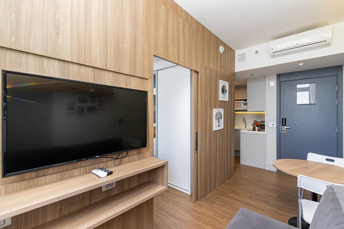 Une chambre moderne équipée d’un téléviseur à écran plat posé sur un meuble en bois, d’une petite table avec des chaises, d’une porte donnant sur une cuisine et d’un climatiseur fixé au mur.