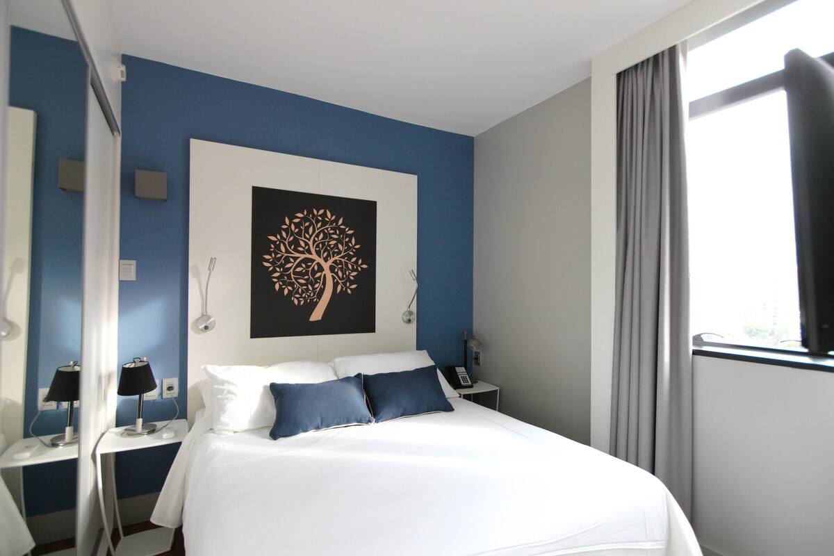 Une chambre d’hôtel avec un lit, deux tables de chevet, une œuvre d’art accrochée au mur, une fenêtre avec des rideaux et une vue sur l’extérieur.