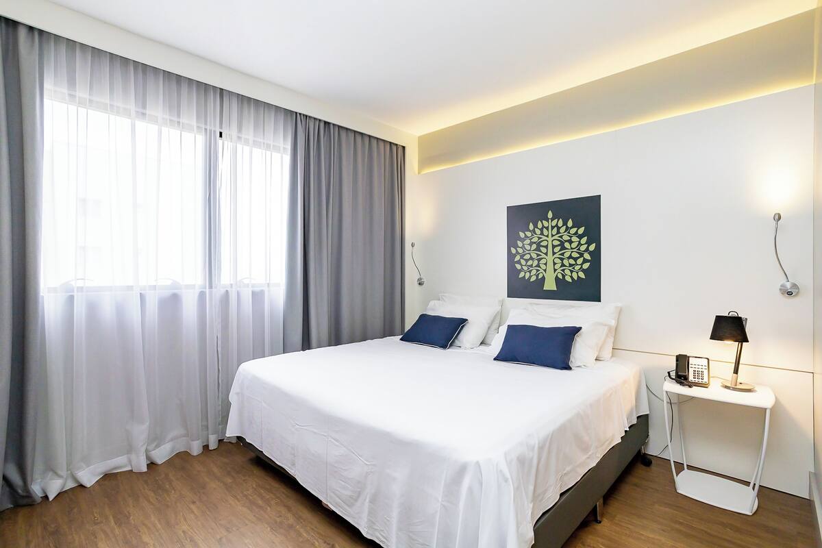 Une chambre d’hôtel avec un grand lit, une table de chevet avec une lampe, un téléphone fixé au mur et une fenêtre avec des rideaux légers.