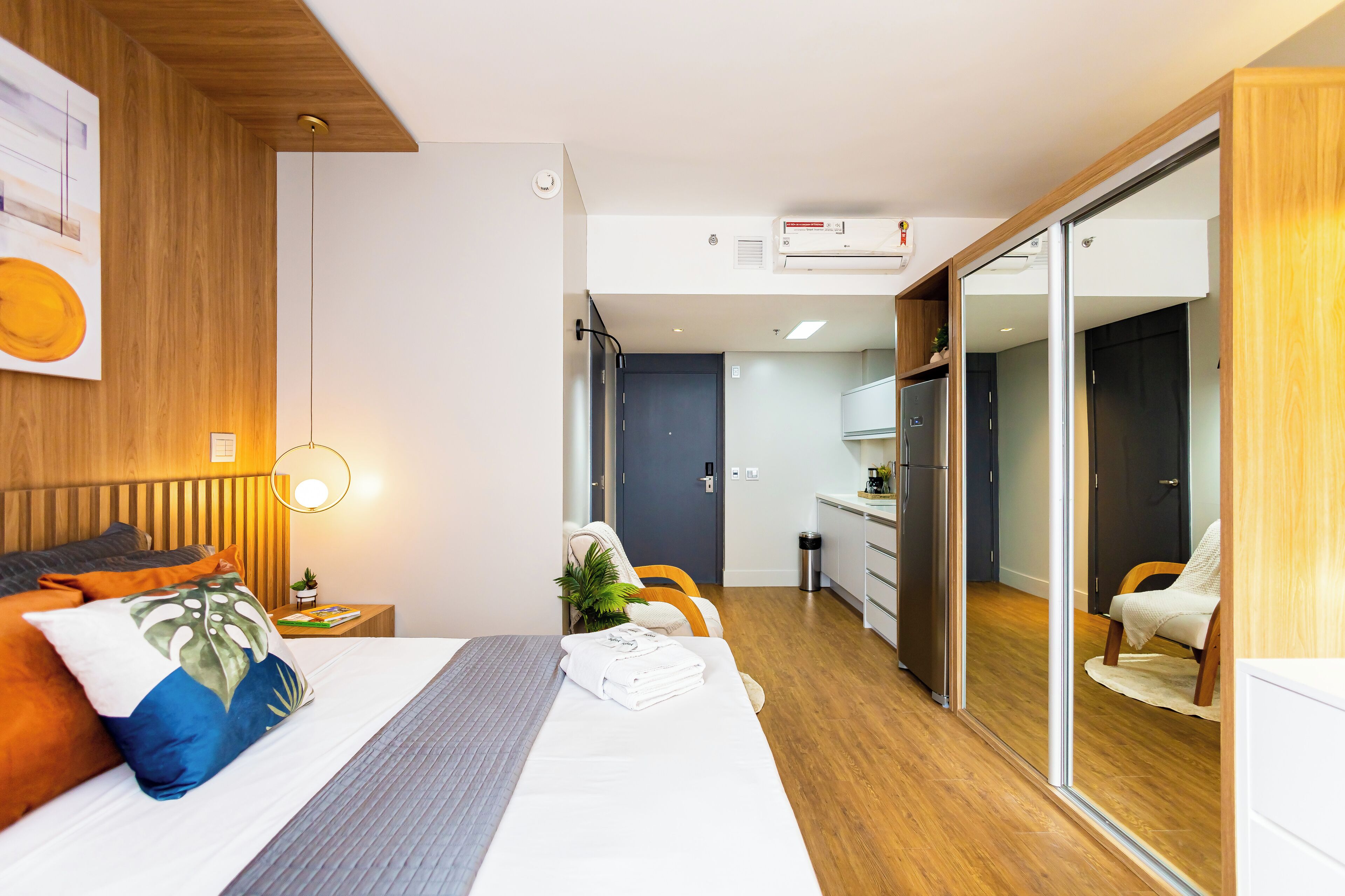 Una habitación de hotel moderna con techo de madera, una cama con cabecera gris, una mesita de noche con lámpara, una planta pequeña y una puerta corredera que da a una kitchenette.