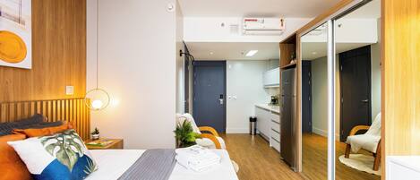 A modern hotel room with a wooden ceiling, a bed with a grey headboard, a bedside table with a lamp, a small plant, and a sliding door leading to a kitchenette.