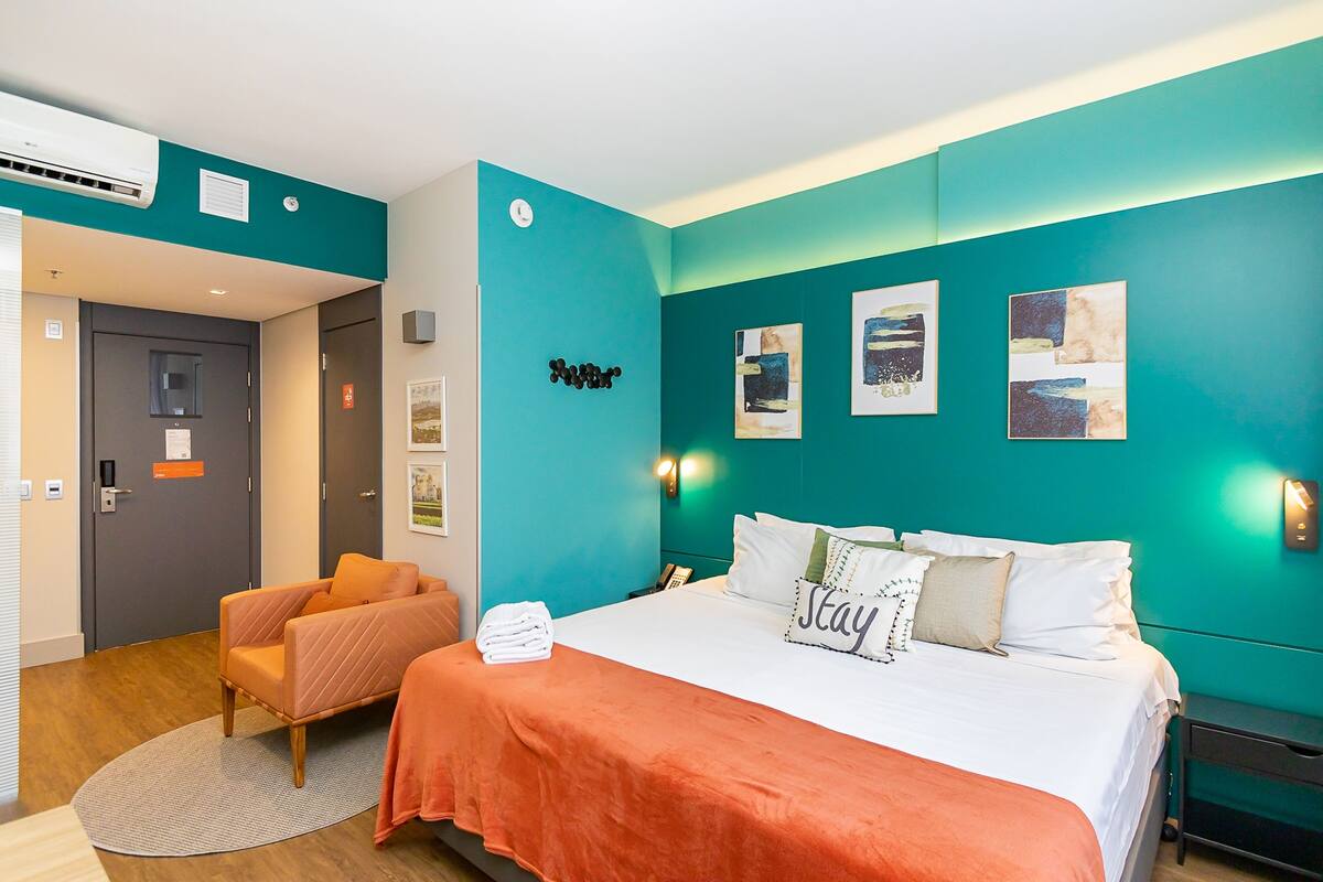 Une chambre d’hôtel aux murs de couleur turquoise, un lit avec du linge de maison blanc, une tête de lit turquoise, un petit fauteuil orange et un tapis rond.
