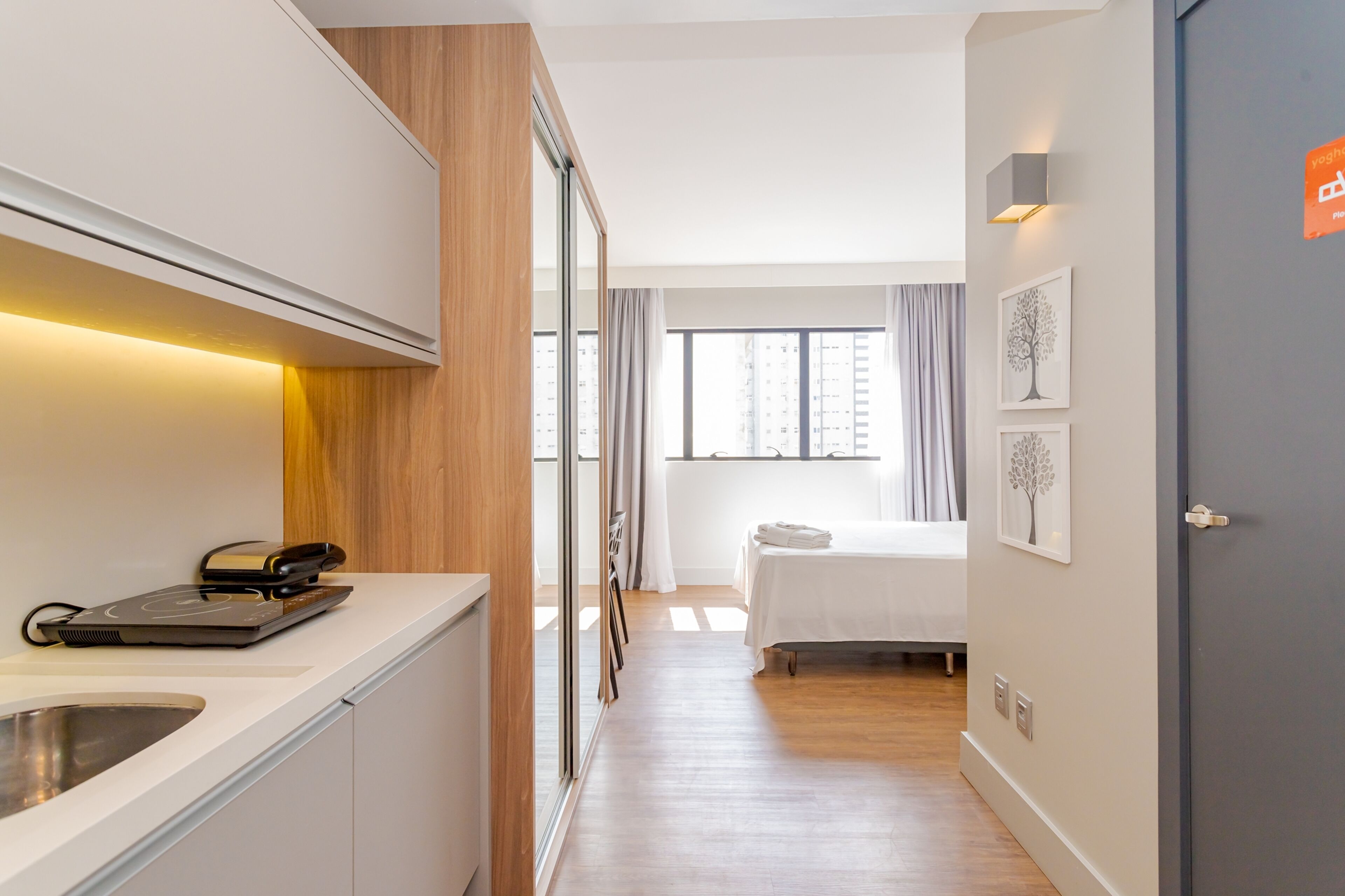 Una habitación de hotel moderna con kitchenette, una cama, un ventanal con cortinas y obras de arte en la pared.